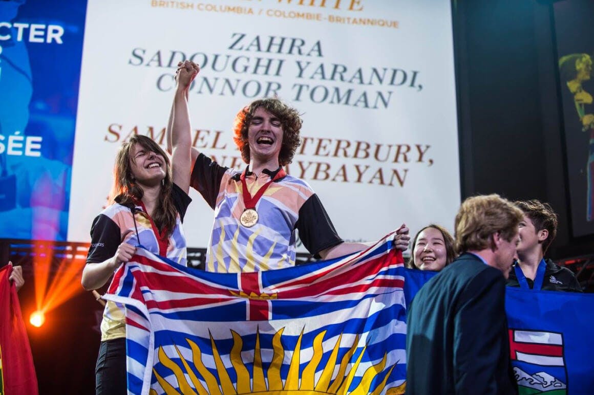 Event photo of students and teachers from the province of British Columbia, showing their team spirit by holding up their provincial flags. They are seen marching towards the main stage during the opening ceremony.